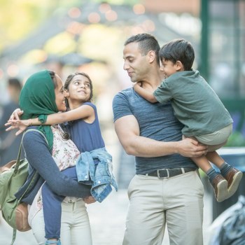 A cute little muslim family smiles at each other. The parents are carrying their kids and the kids have their arms wrapped around their parents.