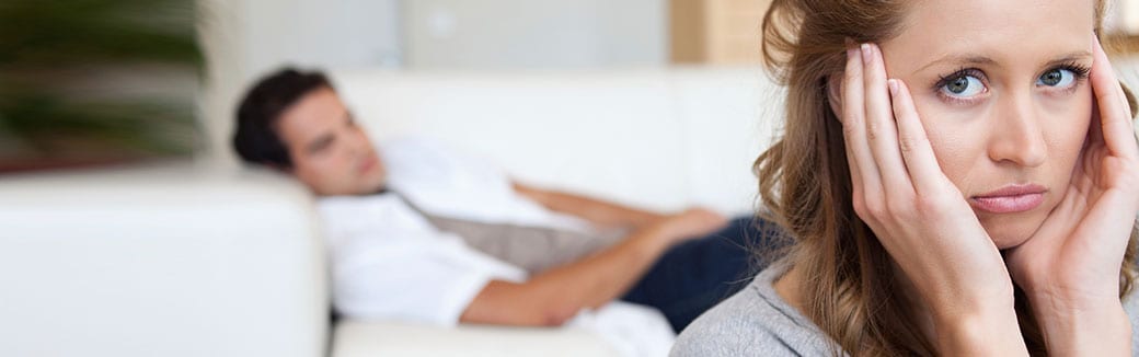 wife looking worried while husband, lying on the couch, looks at her in the background.
