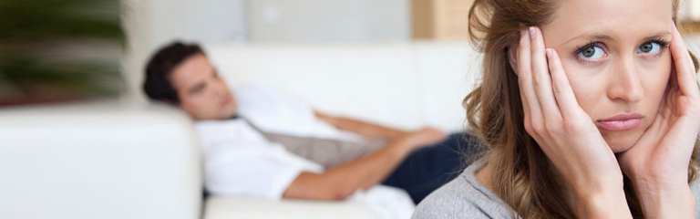 wife looking worried while husband, lying on the couch, looks at her in the background.
