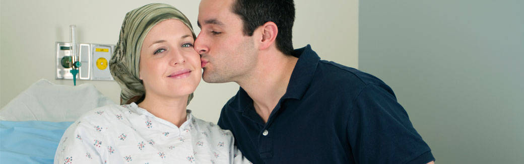 husband kissing his wife on the cheek as she sits in a hospital bed with her head wrapped in a scarf.