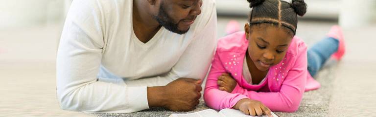 father and daughter laying on their stomachs on the floor looking at a book together.