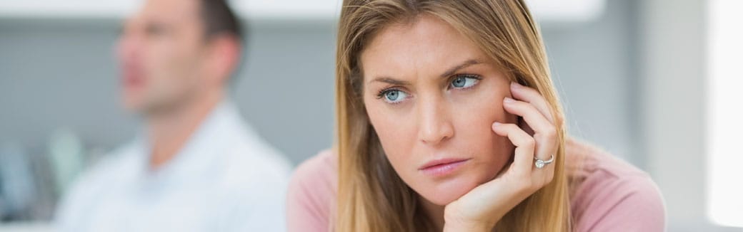 wife looking annoyed with her chin leaning on her hand, and her husband sitting behind her looking away.