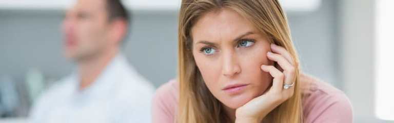 wife looking annoyed with her chin leaning on her hand, and her husband sitting behind her looking away.
